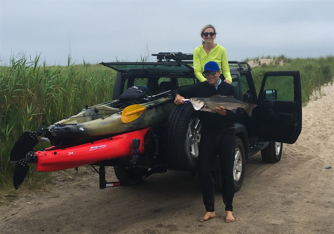 An angler holding a striped bass beside a Jeep loaded with two fishing kayaks on a sandy beach road